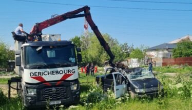 Rescue crane lifting a damaged black car in a grassy lot with onlookers and a blue sky behind.