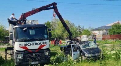 Rescue crane lifting a damaged black car in a grassy lot with onlookers and a blue sky behind.