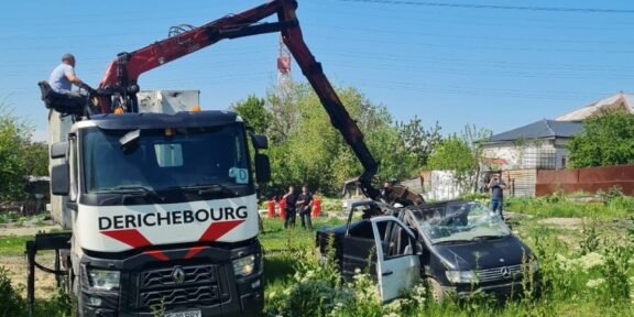 Rescue crane lifting a damaged black car in a grassy lot with onlookers and a blue sky behind.