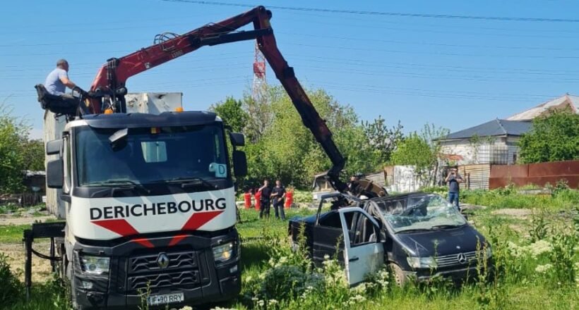 Rescue crane lifting a damaged black car in a grassy lot with onlookers and a blue sky behind.