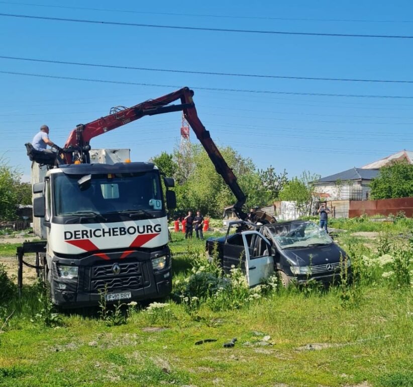 Rescue crane lifting a damaged black car in a grassy lot with onlookers and a blue sky behind.