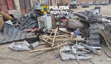 Pile of scrap metal, wood, and debris scattered on a street with a police logo overlaid and parked cars in the background, indicating a cleanup scene.
