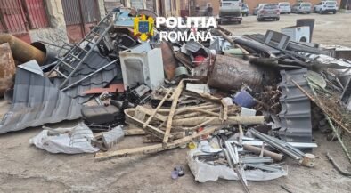 Pile of scrap metal, wood, and debris scattered on a street with a police logo overlaid and parked cars in the background, indicating a cleanup scene.