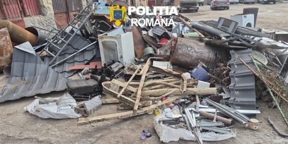 Pile of scrap metal, wood, and debris scattered on a street with a police logo overlaid and parked cars in the background, indicating a cleanup scene.