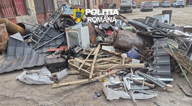 Pile of scrap metal, wood, and debris scattered on a street with a police logo overlaid and parked cars in the background, indicating a cleanup scene.
