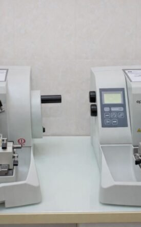 Two compact gray laboratory instruments with control panels sit side by side on a lab bench, each with a sample clamp and cutting area.