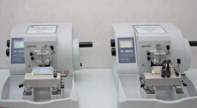 Two compact gray laboratory instruments with control panels sit side by side on a lab bench, each with a sample clamp and cutting area.