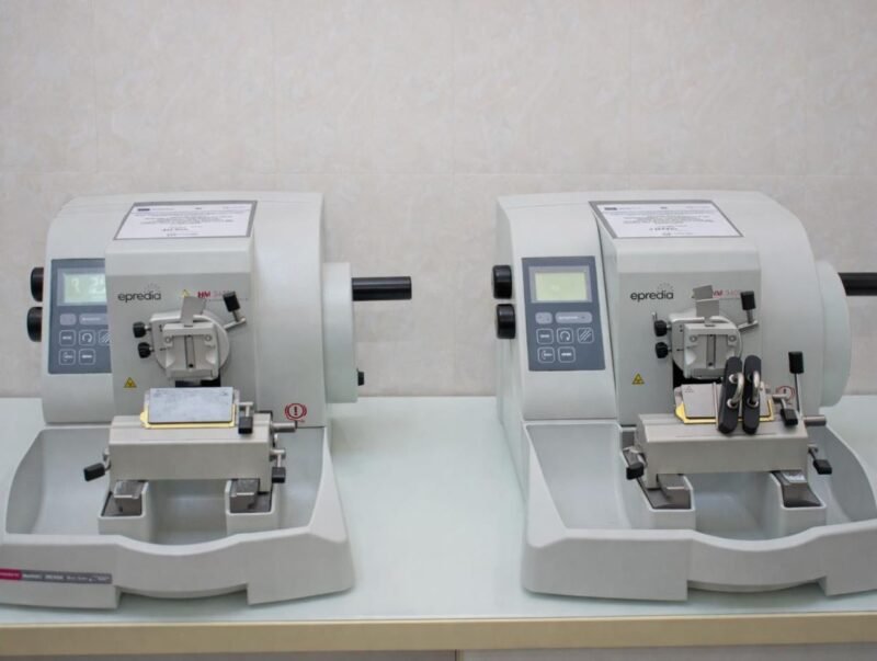 Two compact gray laboratory instruments with control panels sit side by side on a lab bench, each with a sample clamp and cutting area.