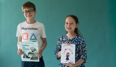 Two smiling children, a boy with glasses in a white PlayStation t-shirt and a girl in a blue floral dress, hold books in front of a teal wall.