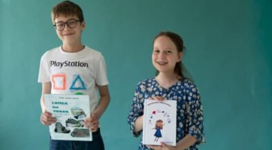 Two smiling children, a boy with glasses in a white PlayStation t-shirt and a girl in a blue floral dress, hold books in front of a teal wall.