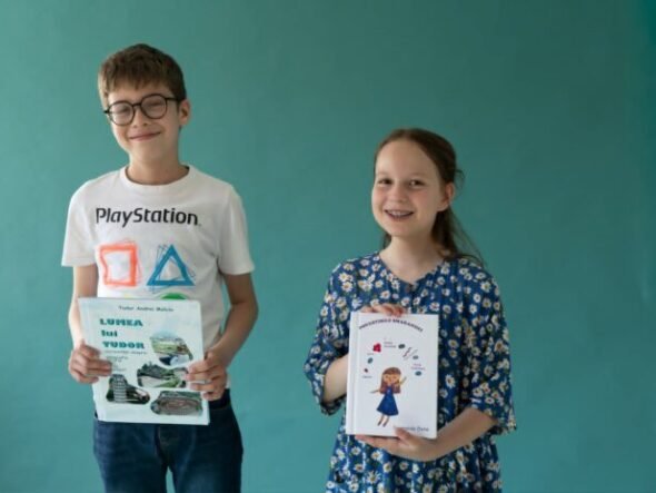 Two smiling children, a boy with glasses in a white PlayStation t-shirt and a girl in a blue floral dress, hold books in front of a teal wall.