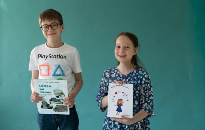Two smiling children, a boy with glasses in a white PlayStation t-shirt and a girl in a blue floral dress, hold books in front of a teal wall.