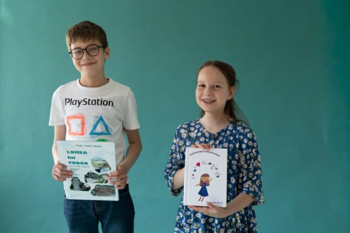 Two smiling children, a boy with glasses in a white PlayStation t-shirt and a girl in a blue floral dress, hold books in front of a teal wall.