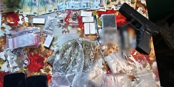 Romanian police display confiscated items on a table: euro banknotes, small bags of drugs, a handgun, and several smartphones.
