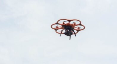 Orange hexacopter drone with six propeller arms hovering in a pale blue sky