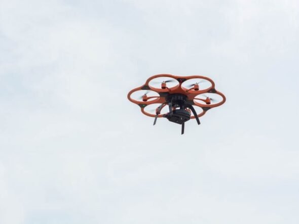 Orange hexacopter drone with six propeller arms hovering in a pale blue sky