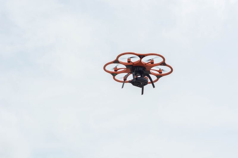 Orange hexacopter drone with six propeller arms hovering in a pale blue sky