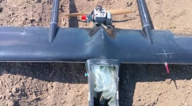 Top-down view of a small single-engine airplane model with a wooden propeller, resting on bare dirt; cockpit area is covered with a plastic bag.