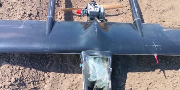 Top-down view of a small single-engine airplane model with a wooden propeller, resting on bare dirt; cockpit area is covered with a plastic bag.