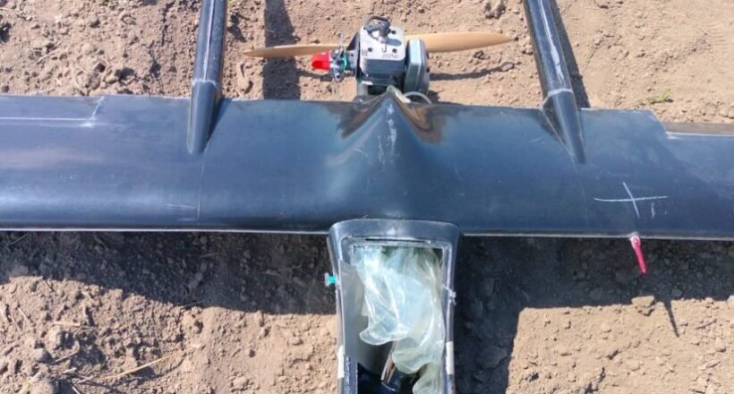 Top-down view of a small single-engine airplane model with a wooden propeller, resting on bare dirt; cockpit area is covered with a plastic bag.