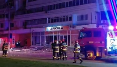 Nighttime scene with firefighters in gear near a red fire truck, blue and red emergency lights, in front of a building labeled 'APICOLA' and debris on the sidewalk.
