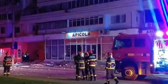 Nighttime scene with firefighters in gear near a red fire truck, blue and red emergency lights, in front of a building labeled 'APICOLA' and debris on the sidewalk.