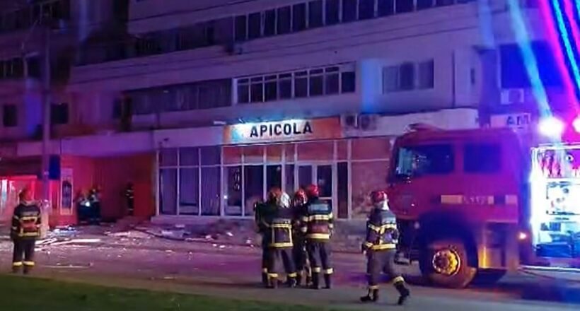 Nighttime scene with firefighters in gear near a red fire truck, blue and red emergency lights, in front of a building labeled 'APICOLA' and debris on the sidewalk.