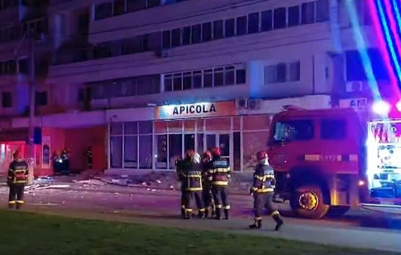 Nighttime scene with firefighters in gear near a red fire truck, blue and red emergency lights, in front of a building labeled 'APICOLA' and debris on the sidewalk.