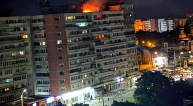 Nighttime view of a long apartment building with a fire on the roof and bright orange flame, street storefronts and cars below.