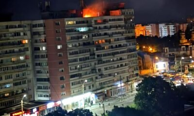 Nighttime view of a long apartment building with a fire on the roof and bright orange flame, street storefronts and cars below.