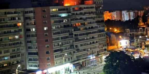 Nighttime view of a long apartment building with a fire on the roof and bright orange flame, street storefronts and cars below.