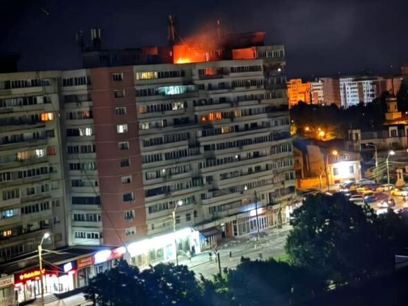 Nighttime view of a long apartment building with a fire on the roof and bright orange flame, street storefronts and cars below.