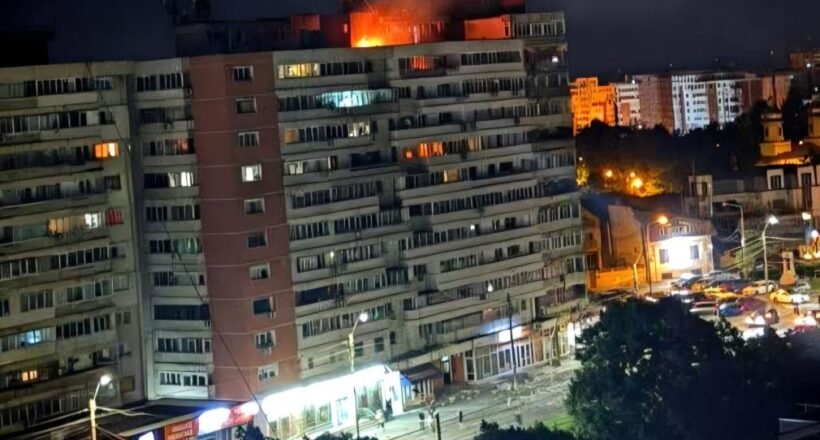 Nighttime view of a long apartment building with a fire on the roof and bright orange flame, street storefronts and cars below.
