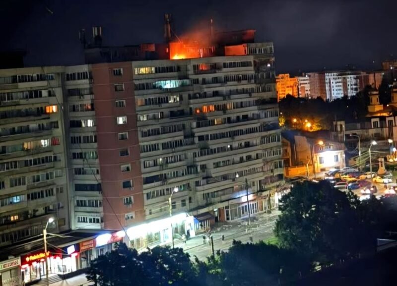 Nighttime view of a long apartment building with a fire on the roof and bright orange flame, street storefronts and cars below.