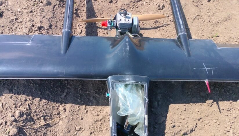 Top-down view of a small single-engine airplane model with a wooden propeller, resting on bare dirt; cockpit area is covered with a plastic bag.