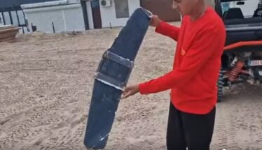 Man in a red shirt holds a large curved blue panel strapped with metal bands in a dusty outdoor yard near a pickup truck.