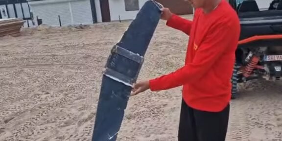 Man in a red shirt holds a large curved blue panel strapped with metal bands in a dusty outdoor yard near a pickup truck.