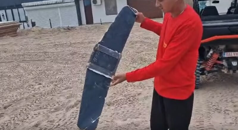 Man in a red shirt holds a large curved blue panel strapped with metal bands in a dusty outdoor yard near a pickup truck.