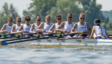Women’s eight rowing team in a white-and-blue boat racing on a lake, each athlete with oars raised and wearing sunglasses and team uniforms.
