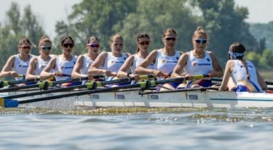 Women’s eight rowing team in a white-and-blue boat racing on a lake, each athlete with oars raised and wearing sunglasses and team uniforms.