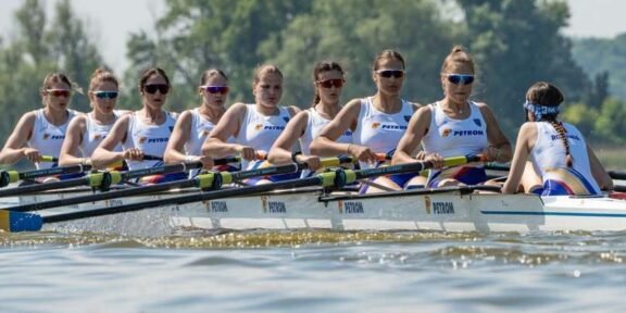 Women’s eight rowing team in a white-and-blue boat racing on a lake, each athlete with oars raised and wearing sunglasses and team uniforms.