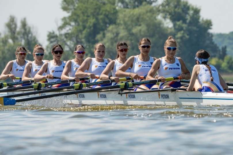Women’s eight rowing team in a white-and-blue boat racing on a lake, each athlete with oars raised and wearing sunglasses and team uniforms.