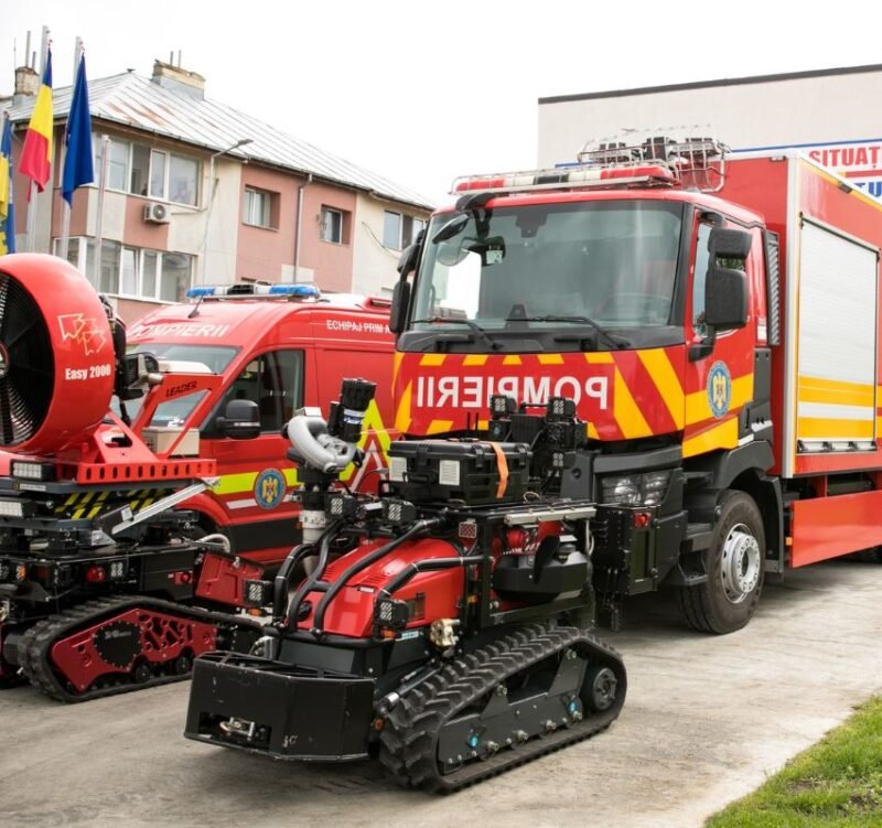 Red fire trucks with yellow chevrons parked in a lot, with a red tracked rescue robot in the foreground.