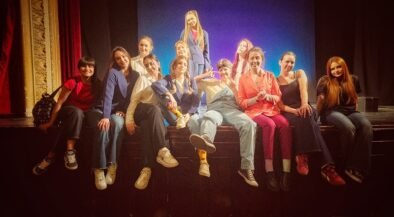 A group of about 12 young people posing together on a theater stage, some sitting on the edge and one standing center, smiling at the camera.