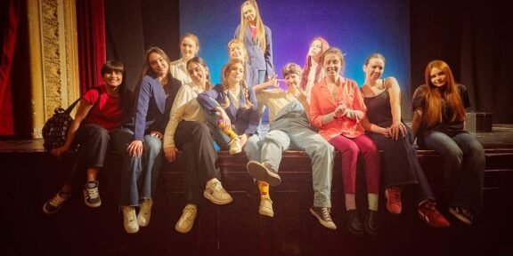 A group of about 12 young people posing together on a theater stage, some sitting on the edge and one standing center, smiling at the camera.