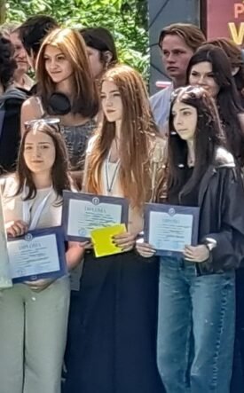 Group of young graduates posing with framed certificates at an outdoor ceremony. A banner is in the background.