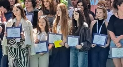 Group of young graduates posing with framed certificates at an outdoor ceremony. A banner is in the background.