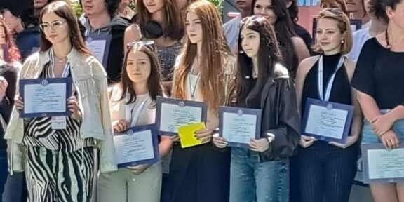 Group of young graduates posing with framed certificates at an outdoor ceremony. A banner is in the background.