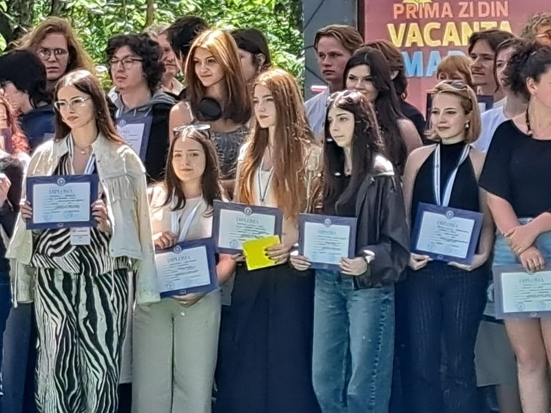 Group of young graduates posing with framed certificates at an outdoor ceremony. A banner is in the background.