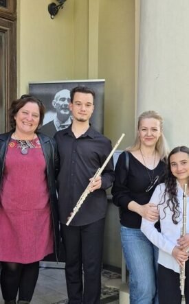 Six people pose on stone steps outside a building, each holding a flute, with a portrait poster in the background.